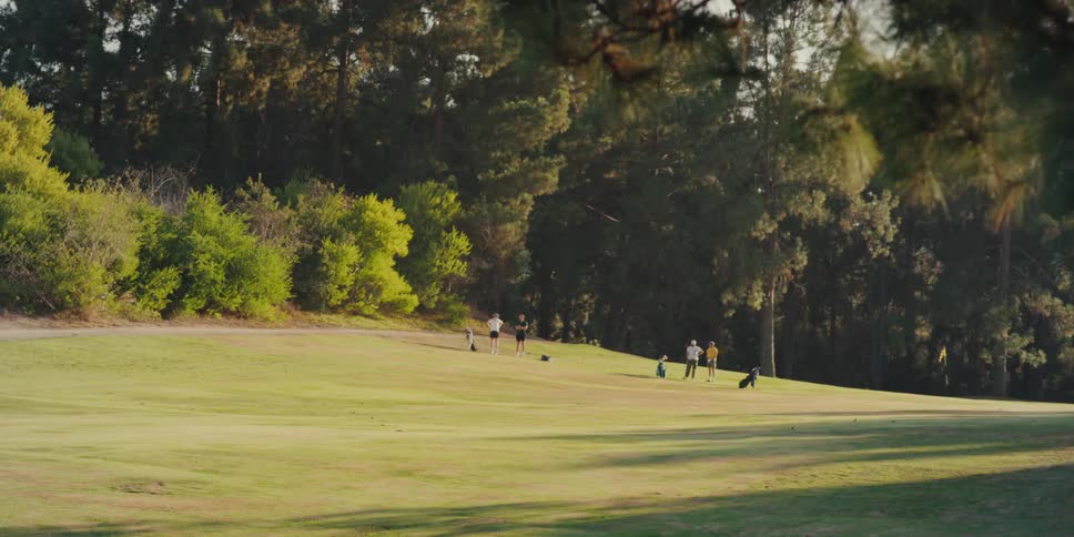 A wide, static shot of people enjoying a sunny day on a beautiful green golf course surrounded by a forest