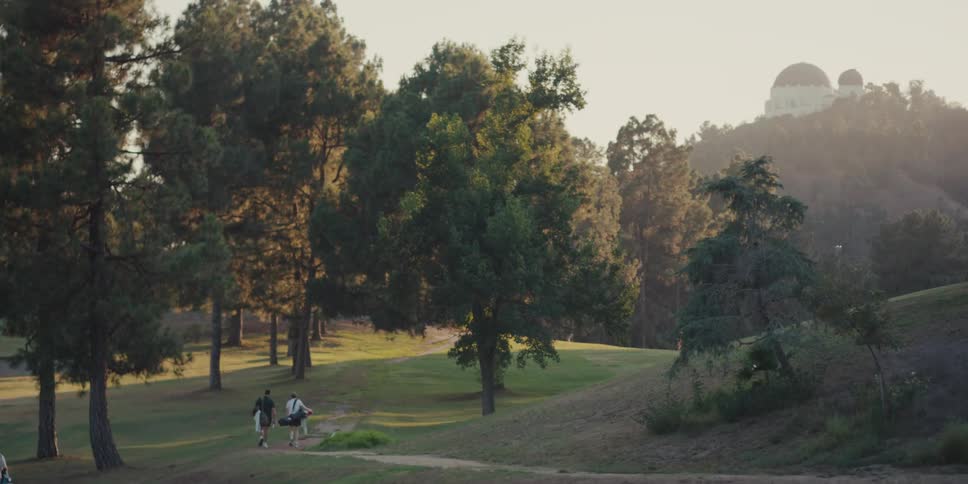 A group of golfers walk up a path on a lush green golf course with the Griffith Observatory visible on a distant hill at sunset
