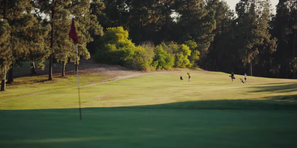 Focus pulls from distant golfers to a red flag on a sun-drenched green during golden hour