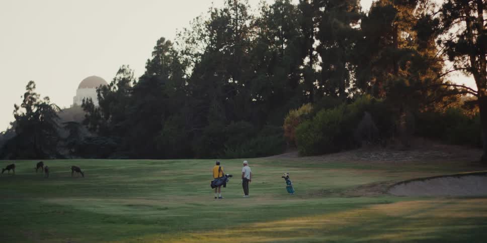 Two golfers enjoy a serene game on a lush course at golden hour as wild deer graze nearby with the Griffith Observatory on the horizon