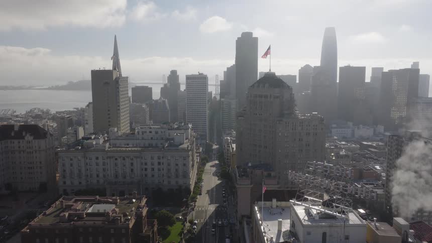 An atmospheric wide shot captures the San Francisco skyline on a hazy day, with steam billowing from a rooftop and the American flag flying