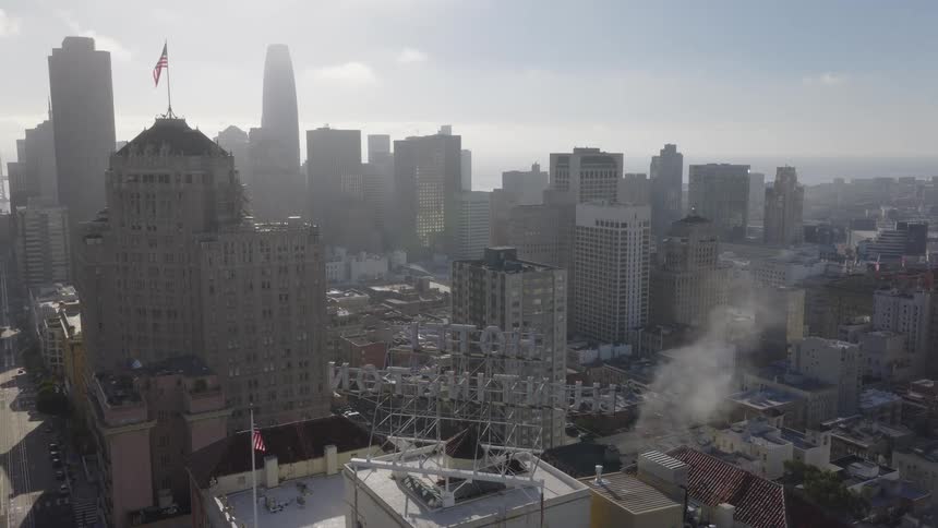 A static high-angle view of the hazy San Francisco skyline, with steam rising from a rooftop and the Huntington Hotel sign in the foreground