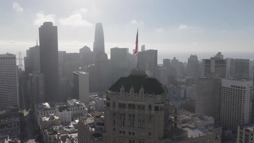 An American flag flies above a historic skyscraper with the iconic San Francisco skyline visible through the morning haze