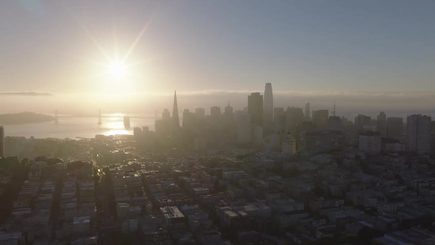 Slow-moving aerial shot reveals the hazy San Francisco skyline and Bay Bridge glowing at sunrise