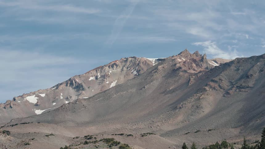 An aerial view slowly approaches the barren, volcanic slopes and snowy peaks of a majestic mountain