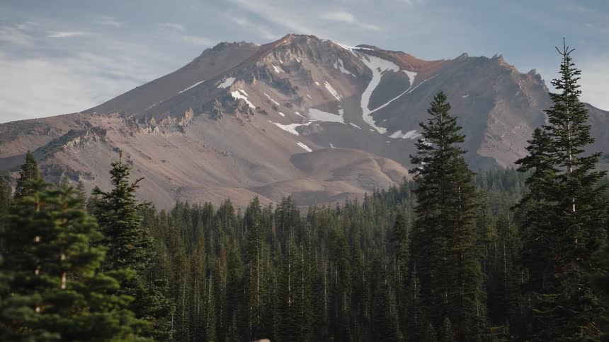 A majestic, snow-dusted mountain towers over a lush, green pine forest under a partly cloudy sky
