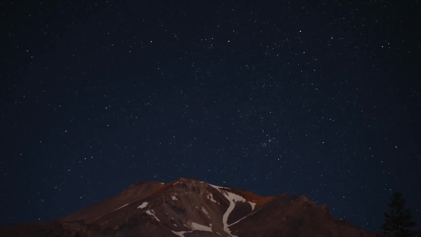 A time-lapse captures satellites streaking across a star-filled night sky over a snow-capped mountain peak