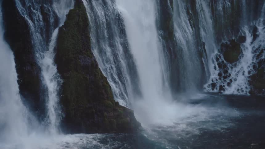 A beautiful close-up shot tilts down a powerful multi-streamed waterfall as it cascades down a mossy cliffside into a churning pool