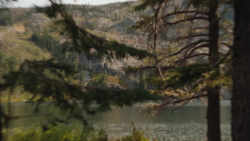 A slow push-in shot reveals a tranquil mountain lake surrounded by a rocky, forested landscape, framed by pine trees in the foreground