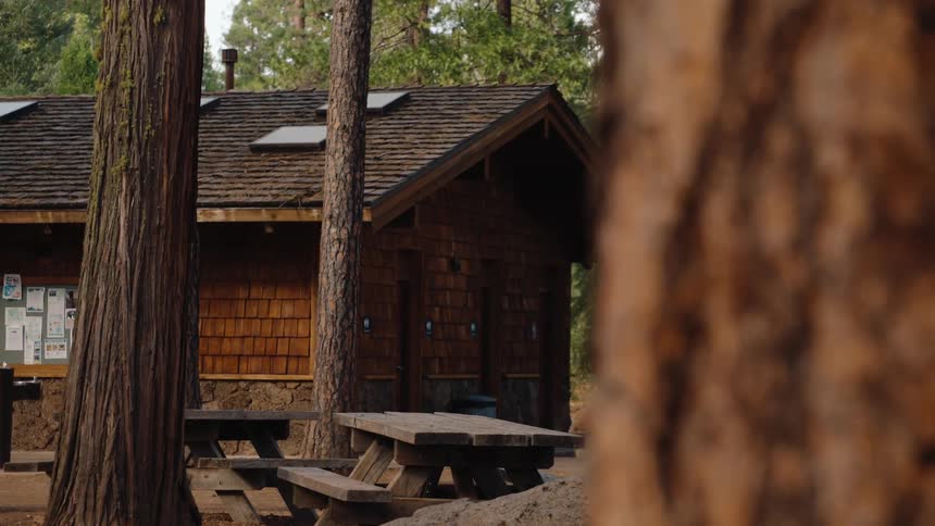 A rustic wooden restroom building with picnic tables at a forest campground with a slow zoom out