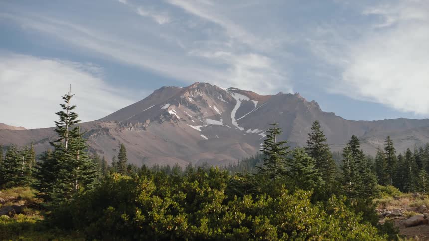 A majestic slow zoom towards the snow-dusted peak of Mount Shasta, towering over a dense evergreen forest on a sunny day