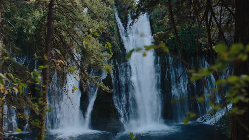 A majestic waterfall cascades down a mossy cliff, viewed through the lush green trees of a forest