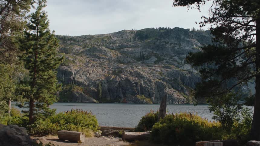 A slow zoom from a forested shoreline across a calm mountain lake reveals the rugged, rocky landscape of the opposite bank