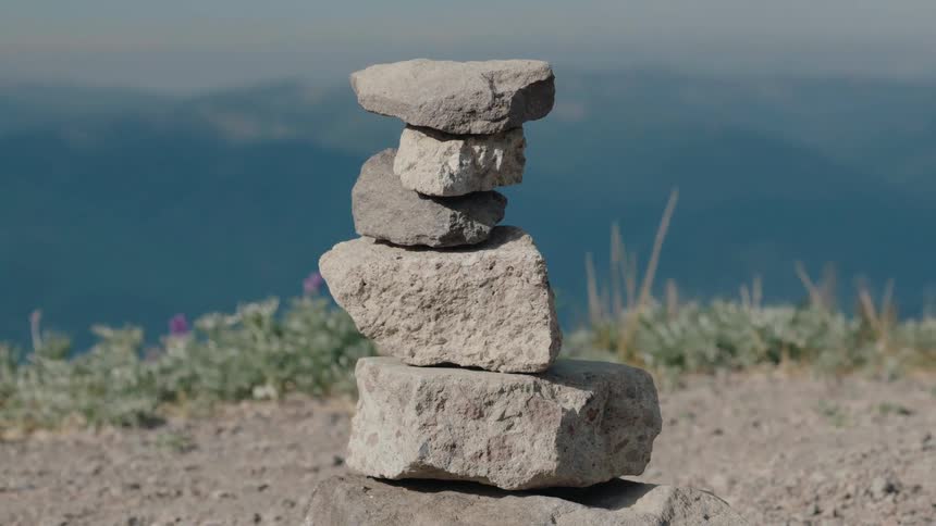 A still close-up of a small, balanced rock cairn on a mountaintop with a hazy, blue mountain range in the distance