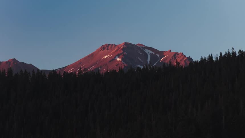 The snow-dusted peak of a majestic mountain is bathed in the warm, pink glow of alpenglow at sunrise above a dark forest