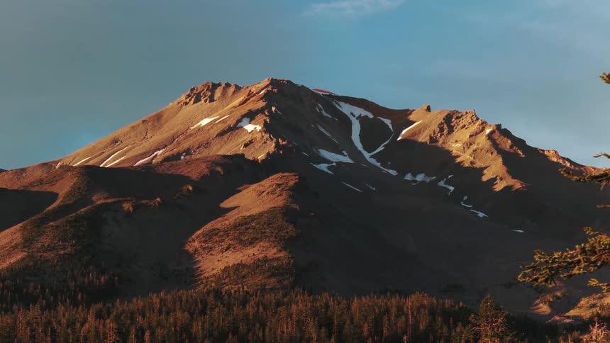 A majestic view of Mount Shasta at golden hour, with warm sunlight highlighting its snow-dusted peaks above a dense pine forest