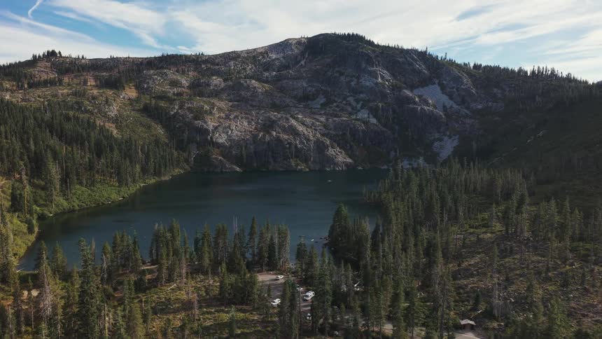A slow aerial push-in over a dense pine forest reveals a secluded alpine lake at the base of a rugged mountain