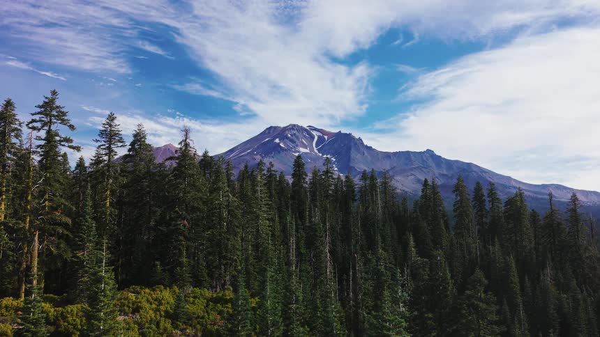 Awe-inspiring aerial view flying over a vast evergreen forest towards the majestic, snow-dusted Mount Shasta under a blue sky with wispy clouds