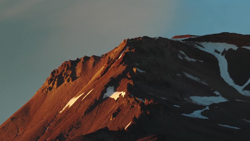 Close-up aerial footage slowly pans across a rocky mountain peak with patches of snow, illuminated by the warm alpenglow of a beautiful sunrise
