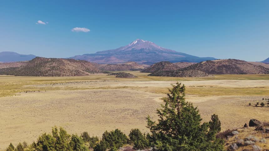 A stunning aerial view reveals the majestic snow-capped Mount Shasta towering over a vast, arid valley on a clear, sunny day