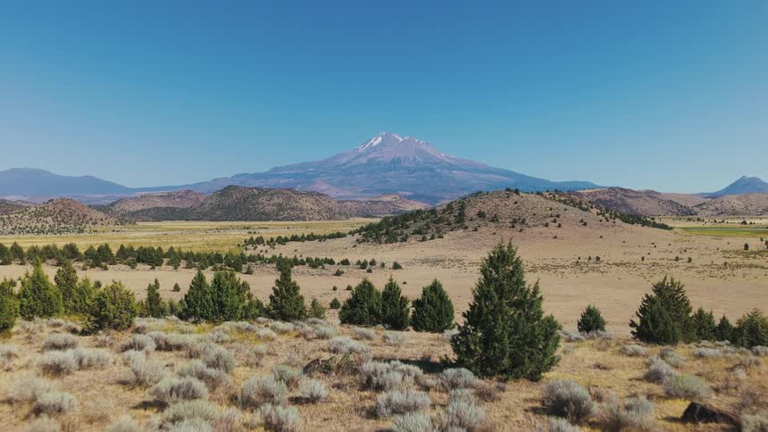 A slow aerial push-in and rise reveals the vast, arid valley landscape with the majestic Mount Shasta prominent on the horizon under a clear blue sky