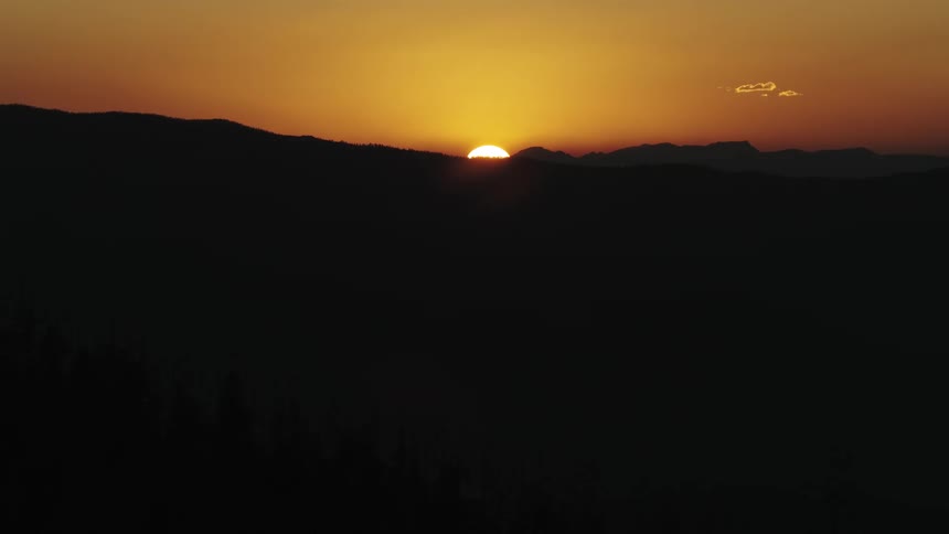 A serene golden sunset dips below the dark silhouette of mountains and a forest