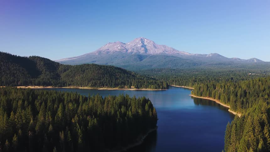 An aerial view reveals the majestic Mount Shasta towering over a pristine blue lake and a lush pine forest on a clear day