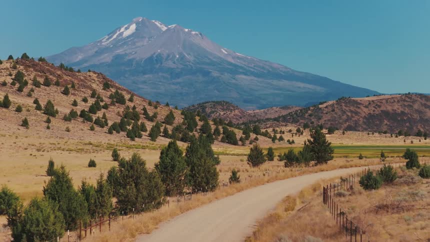 A slow aerial ascent along a winding dirt road reveals the majestic, snow-capped Mount Shasta against a clear blue sky