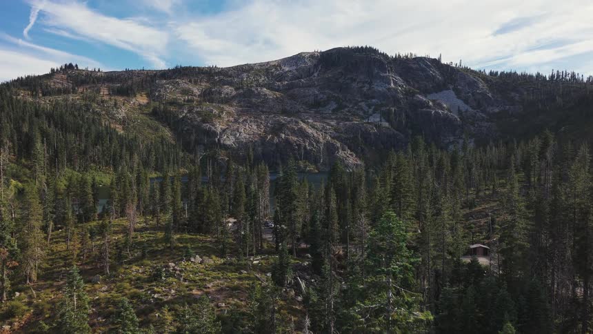 Drone footage reveals a vast, rugged mountain landscape with a pristine alpine lake surrounded by a dense evergreen forest
