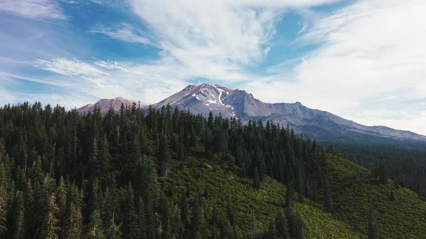 A majestic snow-capped mountain peak rises above a lush green pine forest under a blue sky with wispy clouds