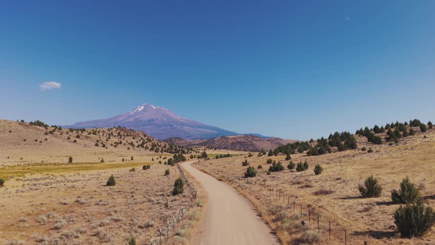 A slow aerial flight travels along a rural dirt road through a vast, arid landscape towards the majestic Mount Shasta on a clear day