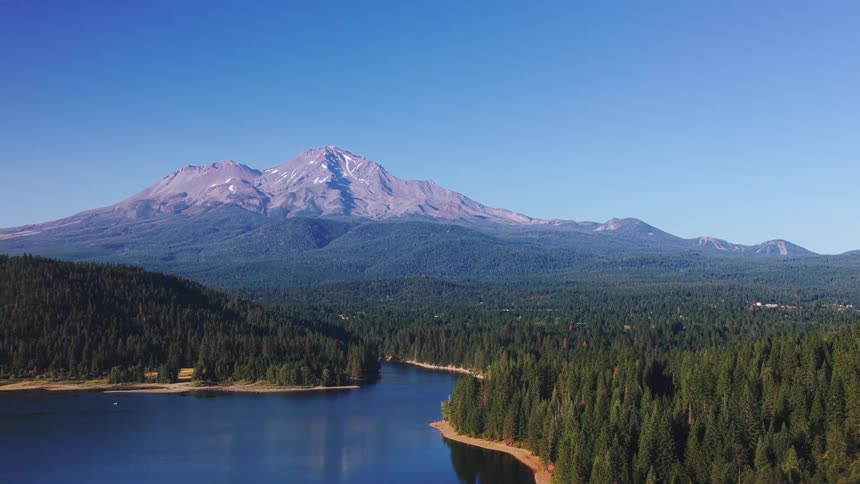 An expansive aerial view reveals the majestic snow-capped Mount Shasta towering over a serene blue lake and a dense evergreen forest