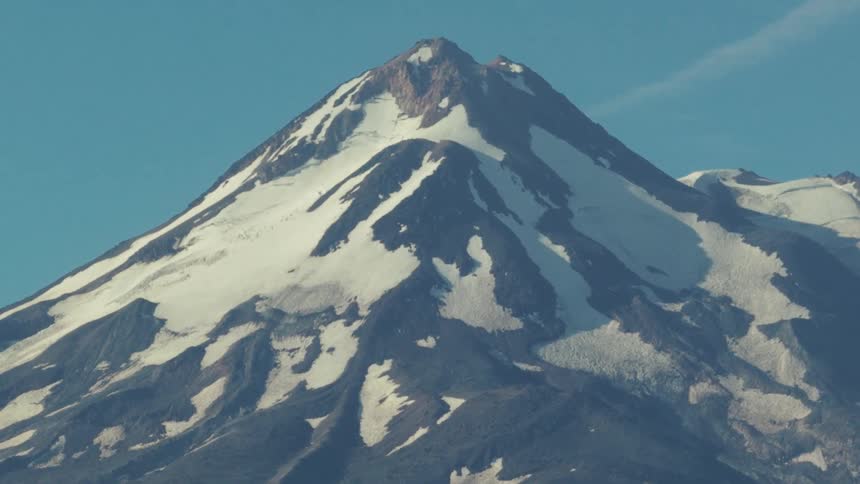 A majestic, snow-covered volcanic peak is revealed in a slow zoom-out against a clear blue sky