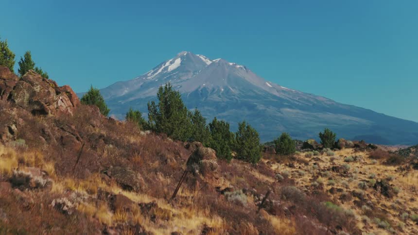 Aerial drone footage rises over a rocky, arid hill to reveal the majestic snow-capped Mount Shasta against a clear blue sky