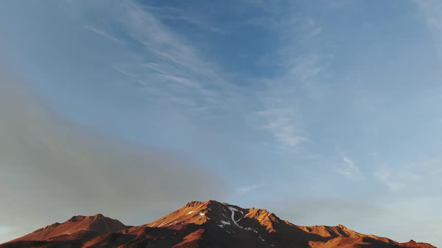 Tilting down from a vast blue sky to reveal a majestic mountain range glowing in the warm light of sunrise