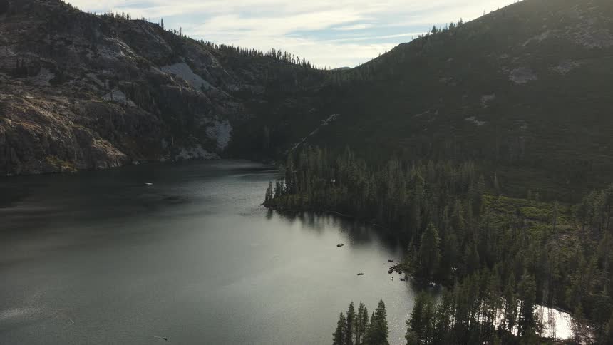 Slow aerial pull-back over a serene alpine lake nestled between rugged mountains and a dense evergreen forest