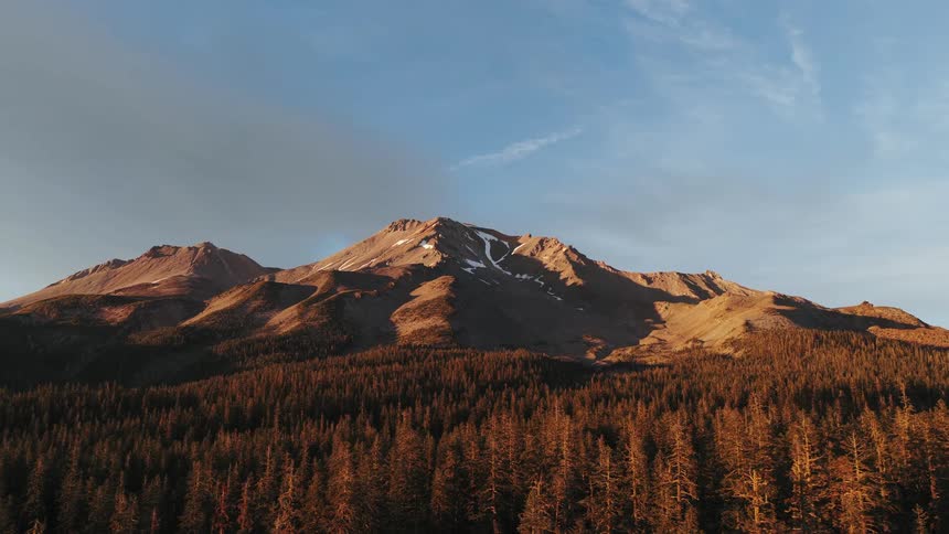 An aerial view tilts down from a serene blue sky to reveal the sun-kissed, snow-capped Mount Shasta rising above a vast evergreen forest at golden hour