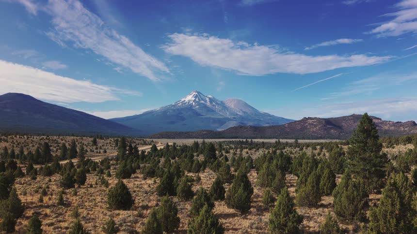 A breathtaking still landscape showcases a snow-capped volcano towering over a high desert forest under a brilliant blue sky