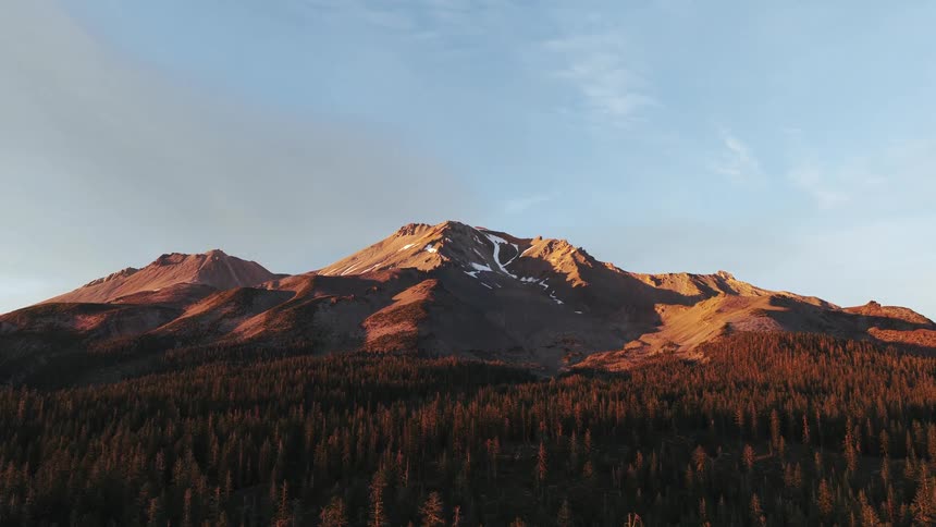 A majestic mountain with snowy peaks glows in the warm golden light of sunset above a dense evergreen forest