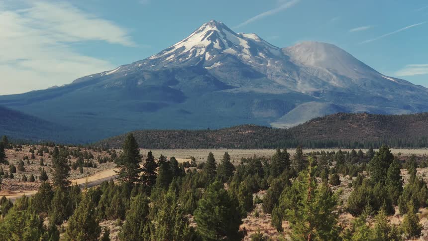 Slow aerial pull-back reveals the majestic snow-capped Mount Shasta towering over a vast, green forest landscape