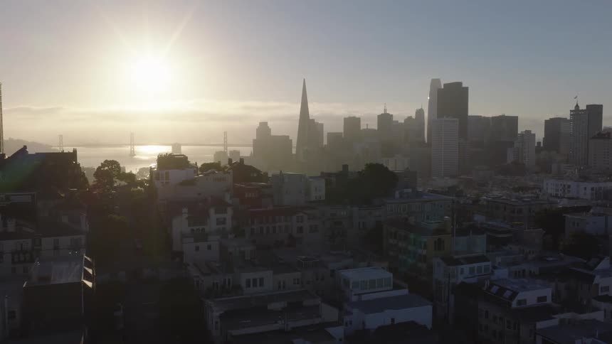 An aerial view of San Francisco shows the city skyline, including the Transamerica Pyramid and Bay Bridge, silhouetted against a bright, hazy sunrise