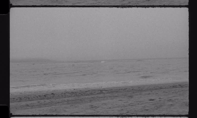 Archival black and white footage of a foggy beach with gentle waves and a bird flying low over the ocean