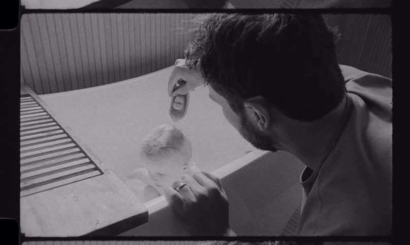A father tenderly bathes his newborn baby in a small bathtub in this intimate, black and white vintage home movie footage