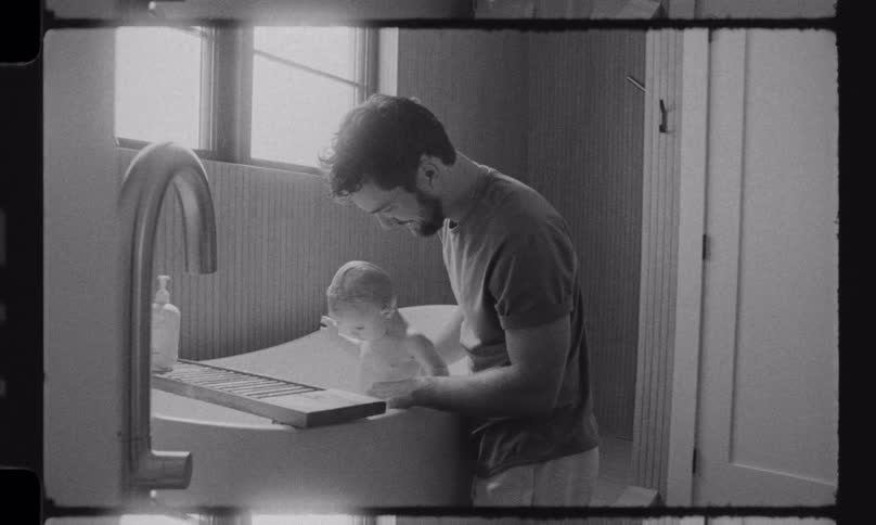 A father gently washes his baby in a freestanding bathtub in this intimate, black and white home movie style clip