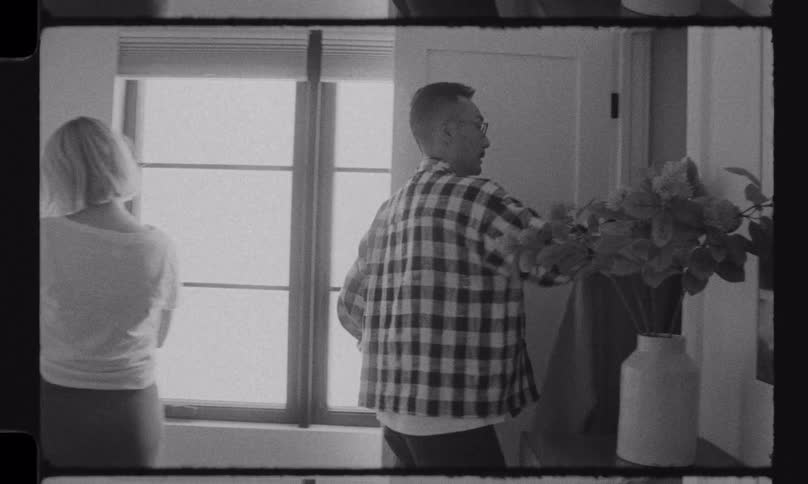 In a grainy, black and white, film-style shot, a man leaves the bedroom while his partner folds laundry alone