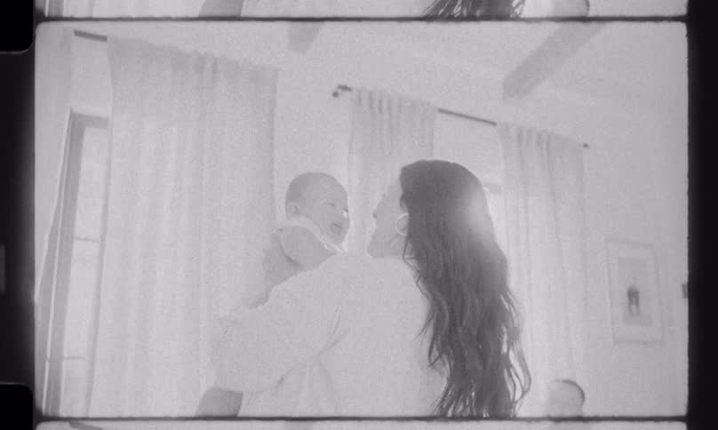 A loving mother playfully lifts her happy baby in a sunlit living room as the father watches from the couch, shot in a nostalgic black and white home movie style
