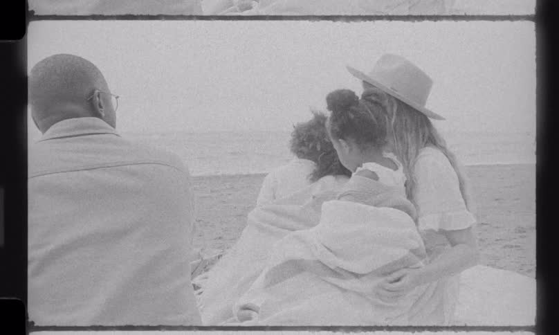 A loving family sits together on a sandy beach looking at the ocean in this nostalgic, black and white vintage footage