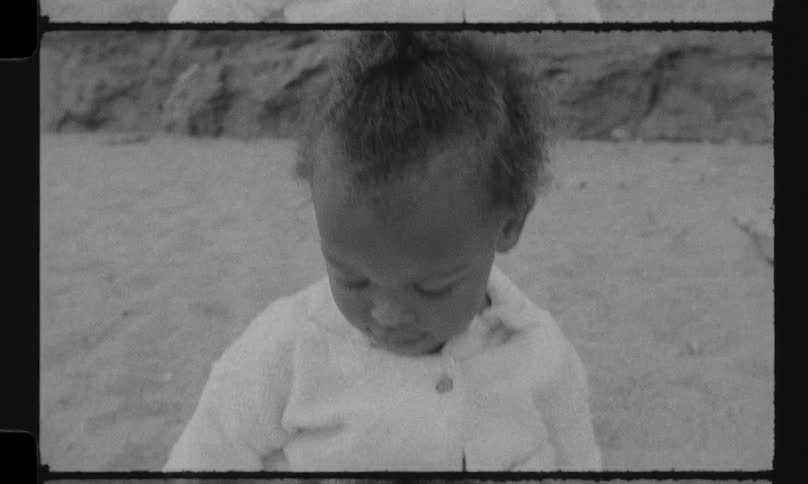A happy African American toddler plays with a stick in the sand at the beach in this vintage black and white film footage