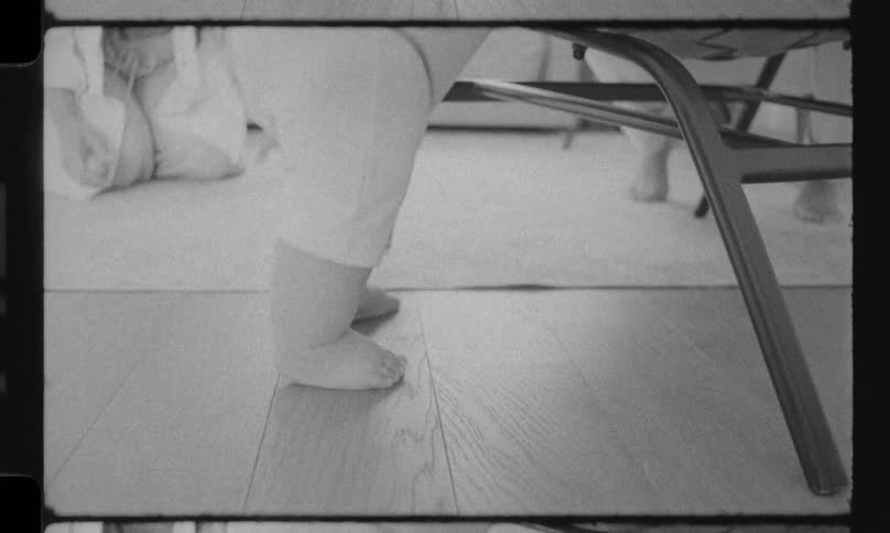 A vintage, black and white shot shows the bare feet of a baby taking its first wobbly steps on a wooden floor at home