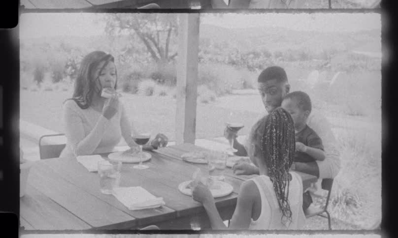 A black and white vintage film shows a happy family of four enjoying a pizza and wine meal together on an outdoor patio in the countryside